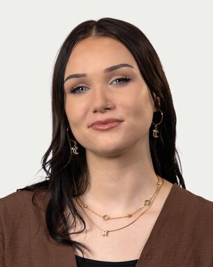 Young woman with long, dark hair, wearing gold earrings and necklaces, looking directly at the camera.