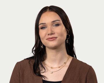 Young woman with long, dark hair, wearing gold earrings and necklaces, looking directly at the camera.