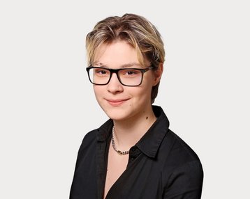 A person with short blonde hair and glasses is wearing a black shirt. The background is neutral and light.