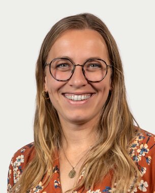 Eine lächelnde Frau mit langen, blonden Haaren und Brille trägt ein blumiges Oberteil. Der Hintergrund ist neutral.