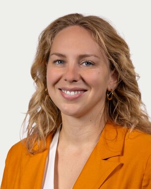 A smiling woman with curly blonde hair is wearing an orange blazer. The background is neutral and bright.