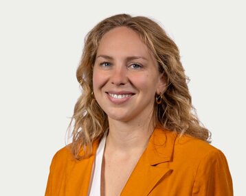 A smiling woman with curly blonde hair is wearing an orange blazer. The background is neutral and bright.