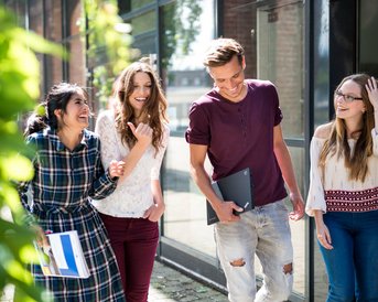 Four young adults are walking together on a sidewalk, smiling and chatting with each other. Two women are dressed in stylish outfits, while the men appear relaxed and are holding notepads.