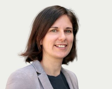 A woman with shoulder-length brown hair and a gray blazer smiles warmly at the camera.