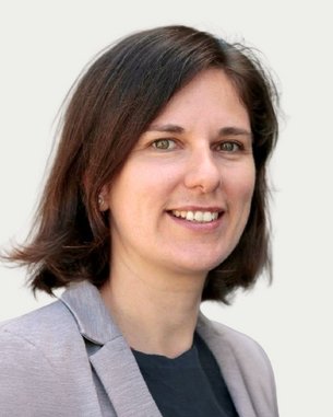 A woman with shoulder-length brown hair and a gray blazer smiles warmly at the camera.