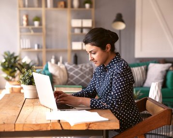 Focused 30s indian business woman  sit at desk in modern cozy home office texting on laptop