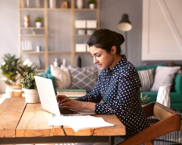 Focused 30s indian business woman  sit at desk in modern cozy home office texting on laptop