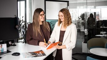 Two women are standing at a desk in a modern office, discussing notes. One woman is holding a pen and writing in a notebook.