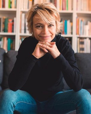 A person with short blonde hair is sitting on a sofa, smiling and resting their chin on their hands. In the background, bookshelves filled with numerous books are visible.