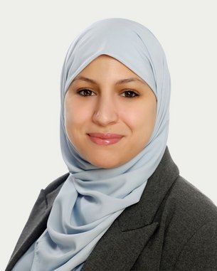 A woman wearing a light blue hijab and a gray blazer smiles at the camera. The background is neutral and bright.