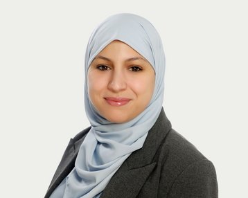 A woman wearing a light blue hijab and a gray blazer smiles at the camera. The background is neutral and bright.