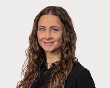 A young woman with curly brown hair and blue eyes is wearing a black top and smiling warmly at the camera.