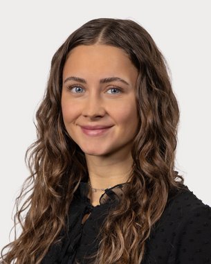 A young woman with curly brown hair and blue eyes is wearing a black top and smiling warmly at the camera.