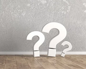 Three large white question marks stand on a wooden floor in front of a gray wall.