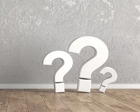 Three large white question marks stand on a wooden floor in front of a gray wall.