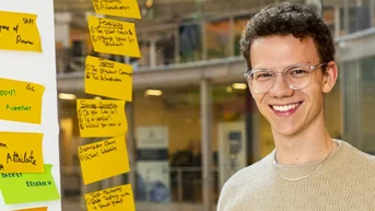 A young man with glasses smiles as he stands at a table, surrounded by colorful sticky notes on a wall.