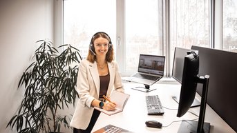 A woman with a headset sits at a desk, working on a computer, surrounded by plants in a modern office.