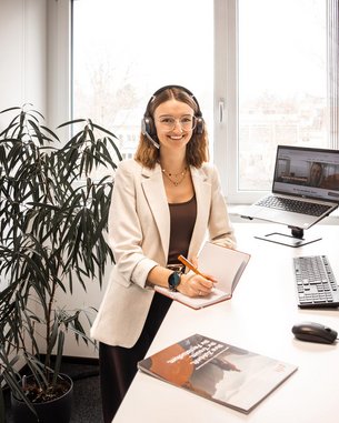 A woman with a headset sits at a desk, working on a computer, surrounded by plants in a modern office.