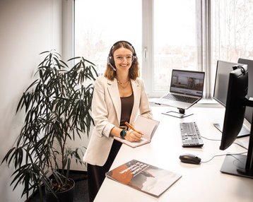 A woman with a headset sits at a desk, working on a computer, surrounded by plants in a modern office.