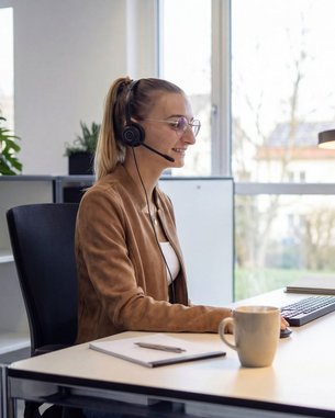 A woman with a headset sits at a desk, working on a computer, surrounded by plants in a modern office.