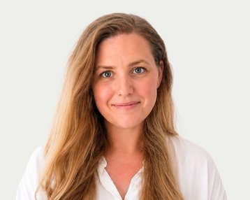 A woman with long, curly hair and a friendly expression is wearing a white shirt. The background is neutral.