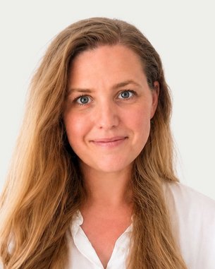 A woman with long, curly hair and a friendly expression is wearing a white shirt. The background is neutral.