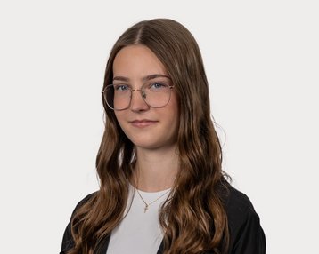 A person with long, curly hair and glasses, standing against a neutral background. They are wearing a black top and have a neutral facial expression.
