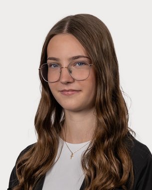 A person with long, curly hair and glasses, standing against a neutral background. They are wearing a black top and have a neutral facial expression.