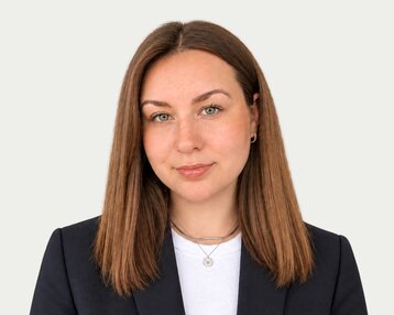 A person with long brown hair and green eyes is wearing a black blazer and a white top.