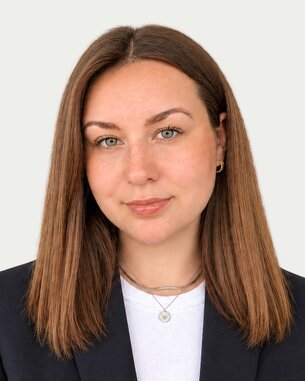 A person with long brown hair and green eyes is wearing a black blazer and a white top.