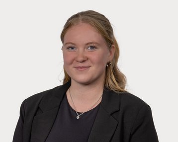 Young woman with blonde hair smiling in a black blazer. The background is neutral and bright.