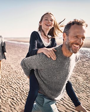 A family is walking along the beach. A man is carrying a girl on his back, while two women and a boy walk nearby. Everyone is smiling and enjoying the day.