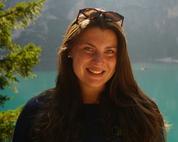 A woman with long brown hair and sunglasses smiles in front of a clear turquoise lake and wooded mountains.