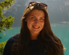 A woman with long brown hair and sunglasses smiles in front of a clear turquoise lake and wooded mountains.