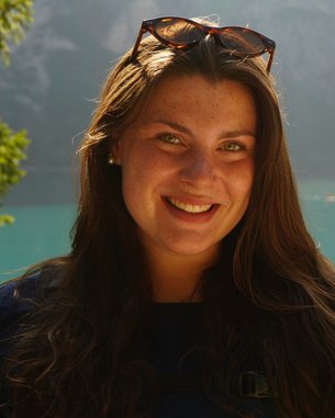 A woman with long brown hair and sunglasses smiles in front of a clear turquoise lake and wooded mountains.