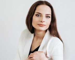 A woman with long brown hair and a neutral expression is wearing a white blazer and posing against a bright background.