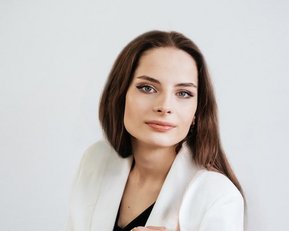 A woman with long brown hair and a neutral expression is wearing a white blazer and posing against a bright background.