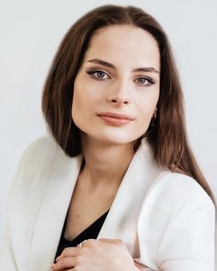 A woman with long brown hair and a neutral expression is wearing a white blazer and posing against a bright background.