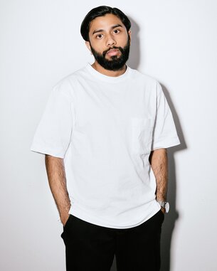 A man with a beard is wearing a white T-shirt and black pants. He stands against a neutral background, with his hands casually in his pockets.