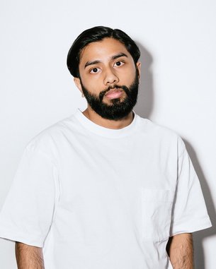 Ein Mann mit Bart trägt ein weißes T-Shirt und schwarze Hose. Er steht vor einem neutralen Hintergrund, die Hände lässig in den Hosentaschen.