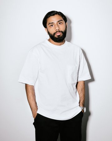 Ein Mann mit Bart trägt ein weißes T-Shirt und schwarze Hose. Er steht vor einem neutralen Hintergrund, die Hände lässig in den Hosentaschen.