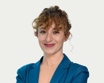 A smiling woman with curly hair is wearing a blue blazer and stands with her arms crossed in front of a bright background.