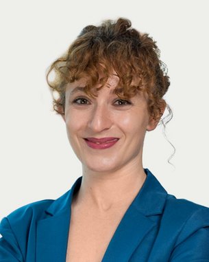 A smiling woman with curly hair is wearing a blue blazer and stands with her arms crossed in front of a bright background.