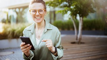 A smiling woman with glasses is sitting on a wooden terrace, holding a smartphone in her hand. She is wearing a light green shirt and has a relaxed posture.