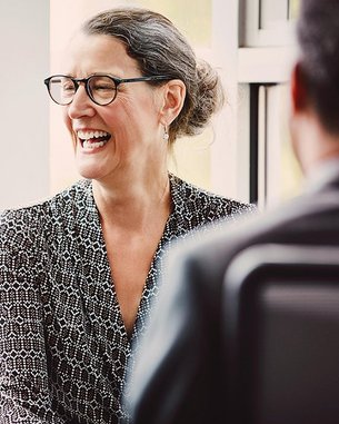 A smiling woman with glasses is wearing a patterned outfit and sitting at a table while talking to another person. The background features windows letting in natural light.