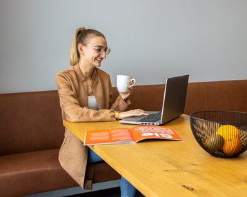 A woman is sitting at a table, working on her laptop and holding a cup in her hand. On the table, there is a magazine and a bowl of fruit.