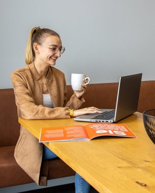 A woman is sitting at a table, working on her laptop and holding a cup in her hand. On the table, there is a magazine and a bowl of fruit.