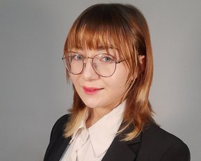A person with shoulder-length brown hair is wearing a black suit and a white blouse. They are standing with their arms crossed, looking directly at the camera.