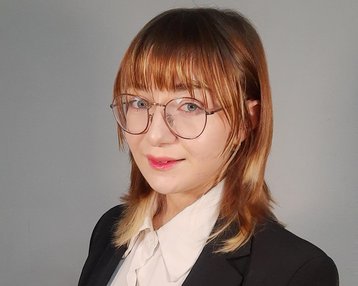 A person with shoulder-length brown hair is wearing a black suit and a white blouse. They are standing with their arms crossed, looking directly at the camera.