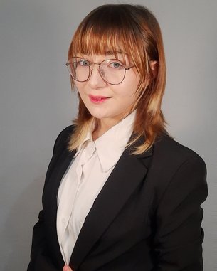 A person with shoulder-length brown hair is wearing a black suit and a white blouse. They are standing with their arms crossed, looking directly at the camera.